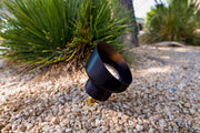 Black outdoor light fixture on a gravel surface with green plants in the background