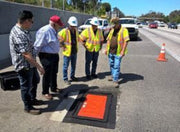 A group of people in high visibility orange vests standing on a black grate inlet protection product, with a focus on the item's size of 48 inches by 48 inches.