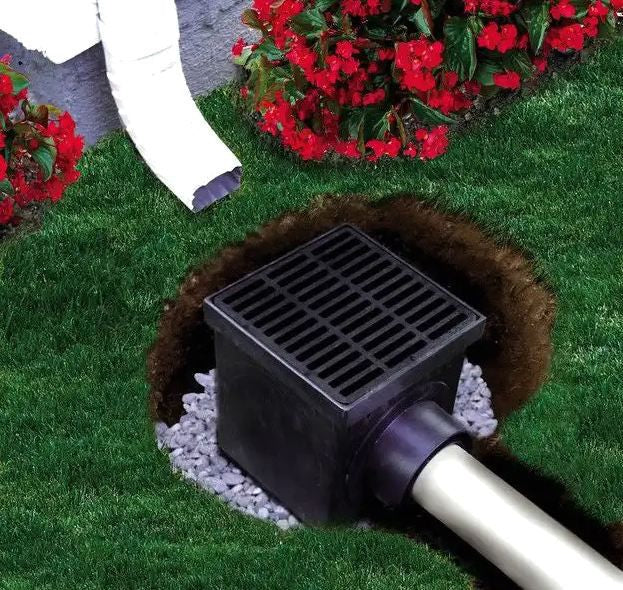 A black catch basin kit with a grate, placed in a garden setting.