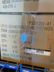 A close-up of a cardboard shipping box for UPVC SCH40 pressure fittings, showing printed product details, a barcode label, and a metal wire rack below.