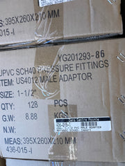 A close-up of stacked brown cardboard shipping boxes with printed black text and a white barcode label. The visible text references 'UPVC SCH40 PRESSURE FITTINGS', 'US4012 MALE ADAPTOR', size 1-1/2", quantity 128 PCS, and measurements 395x260x210 mm.