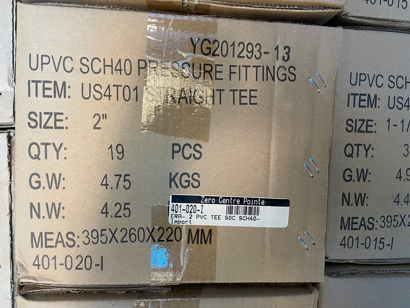 A cardboard shipping box printed with product details for UPVC SCH40 pressure fittings, specifically a straight tee 2-inch size. The box shows printed specifications, a barcode label, and a blue reflective sticker applied to the front.