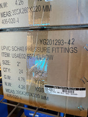 A cardboard shipping box for UPVC SCH40 pressure fittings, elbow, size 2 inches, with printed specifications, a barcode label, and tape on a metal rack.