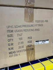 A close-up of a brown cardboard shipping box printed with product details for UPVC SCH40 pressure fittings, item US4005 reducing ring, size 3"x2", quantity 100 pieces, with a barcode label on a yellow and blue storage rack.