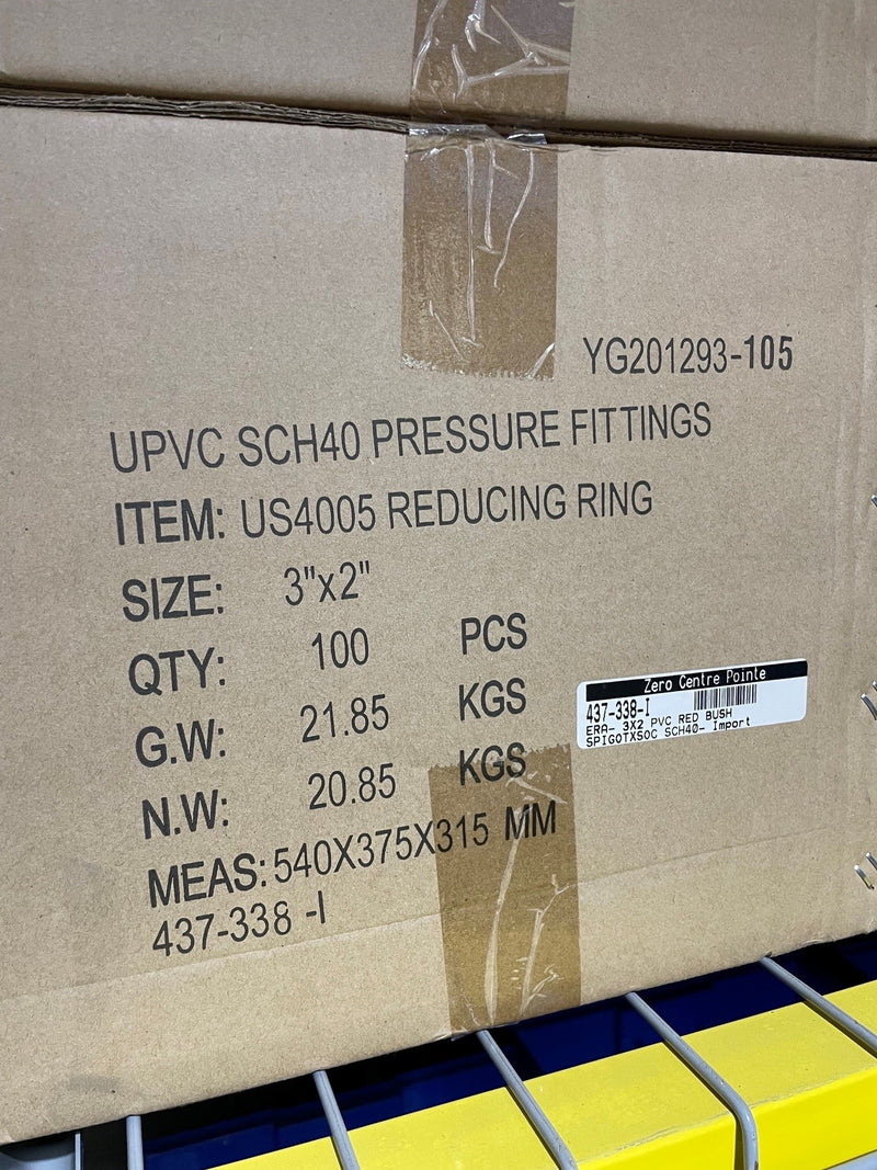 A close-up of a brown cardboard shipping box printed with product details for UPVC SCH40 pressure fittings, item US4005 reducing ring, size 3"x2", quantity 100 pieces, with a barcode label on a yellow and blue storage rack.