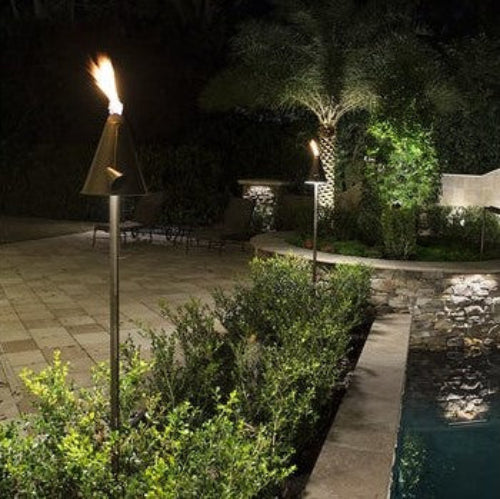 Two polynesian tiki torch lights with bronze-colored metal, placed in a garden setting near a pool at night.