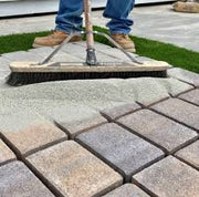 A person is using a tool to apply logic sand between the joints of interlocking concrete pavers that are being laid to create a new surface.