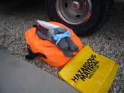 Orange bag with gray items and a yellow 'Hazardous Material' bag on gravel next to a vehicle tire.