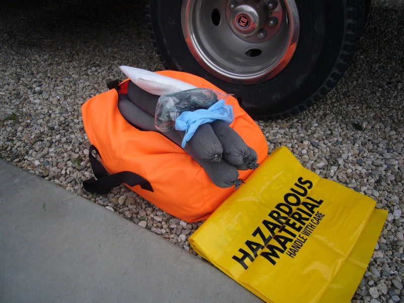 Orange bag with gray items and a yellow 'Hazardous Material' bag on gravel next to a vehicle tire.