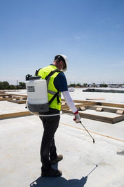 A person wearing a reflective vest and a cap is using a 4 gallon backpack sprayer with a brass nozzle and wand on a construction site.