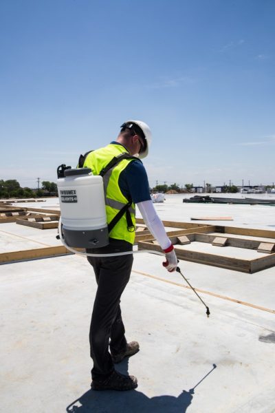 A person wearing a reflective vest and a cap is using a 4 gallon backpack sprayer with a brass nozzle and wand on a construction site.